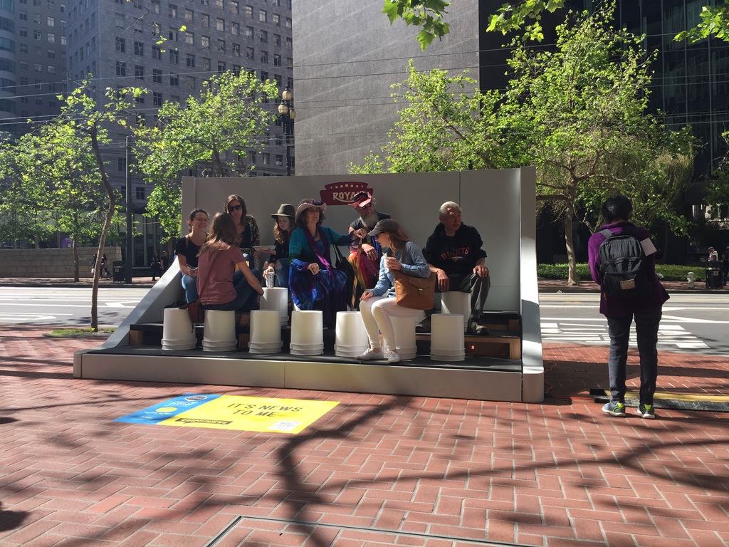 Eight people sitting on the oversized white keys of a giant Royal typewriter installed on a Market Street sidewalk, with office towers and street trees behind.